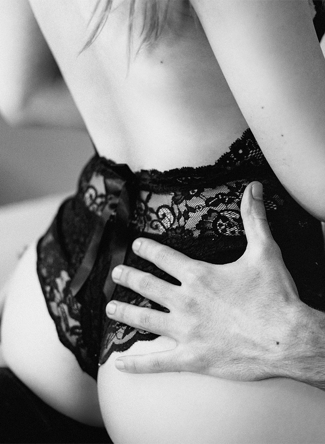 A seductive woman wearing black lace lingerie sits provocatively on a naked man's lap against a black backdrop
